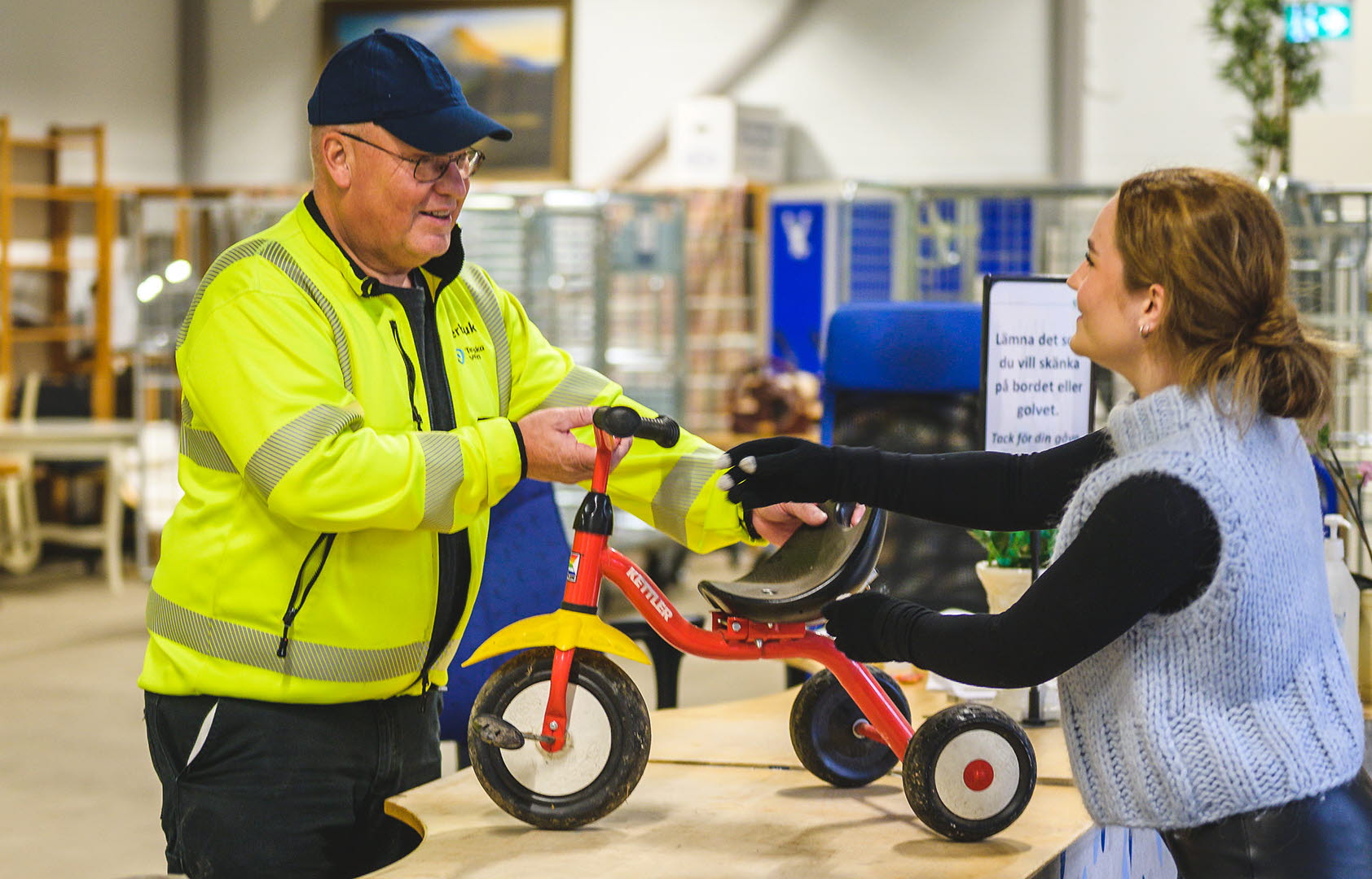 En kvinna lämnar in en liten trehjuling till återbruk, till en medarbetare på Återbrukshallen.